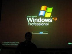 Tantek Çelik stands silhouetted in front of a projection screen on which can be seen a giant Windows XP bootup screen.